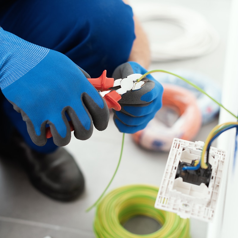 Installation Électricité : Couper un fil avec pince Électricien en gants bleus coupant un fil vert-jaune avec une pince rouge près d'une prise murale nue et de bobines de câble.