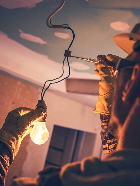 Installation électrique : électricien testant un circuit Électricien avec gants tenant une ampoule allumée tout en testant un fil électrique avec un tournevis testeur au plafond.