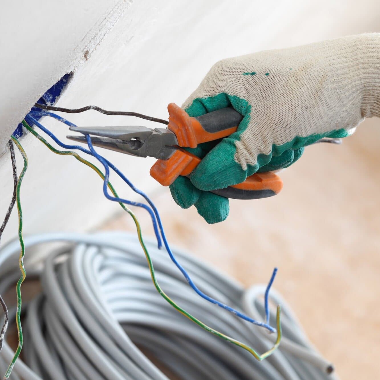 Installation électrique : électricien avec pince et câbles Électricien ganté utilisant une pince à bec effilé pour manipuler des fils électriques colorés sortant d'un mur, avec un rouleau de câble gris en arrière-plan.